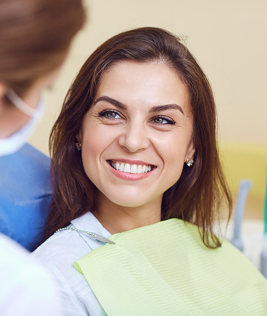 A woman is seated in a dental chair, smiling at the camera, with a dentist wearing a surgical mask and stethoscope attending to her.