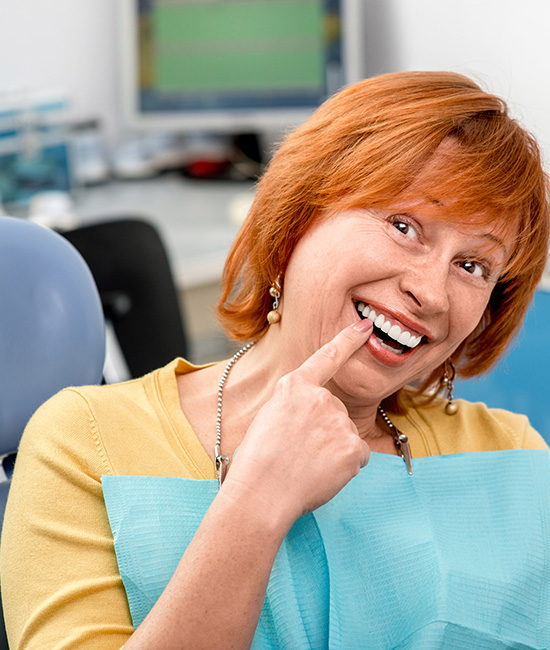 The image is a photograph of an older woman with red hair, wearing glasses and a smile. She has her hand on her chin in a playful manner while sitting at a dental office chair.